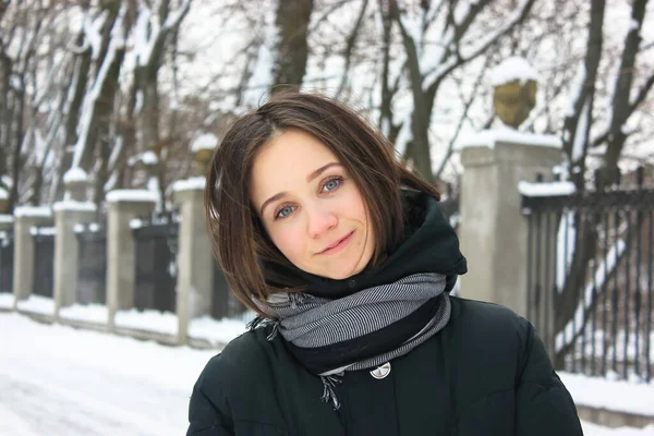 Happy young brunette with a basic bob haircut in a black coat with a scarf. Ukrainian girl smiling standing in a snow-covered winter park in the afternoon. Kiev, Europe.