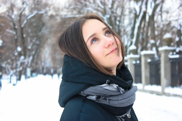 Happy young brunette with a basic bob haircut in a black coat with a scarf. Ukrainian girl smiling and looking in the sky standing in a snow-covered winter park in the afternoon. Kyiv, Europe.