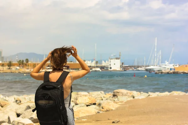 Chica turista de pelo rubio con una mochila negra va a lo largo del paseo marítimo, estacionamiento para yates en el horizonte — Foto de Stock