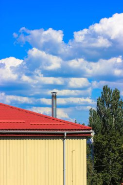 Yazın beyaz bulutlu ve yeşil ağaçlı mavi gökyüzüne karşı kırmızı kiremitli çatılı bir bina.
