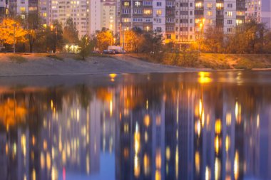 Pencerelerinde parlak ışık olan çok katlı evlerin siluetleri göldeki mavi su yüzeyini yansıtıyor. Geceleri gölün kıyısında geziniyorum.