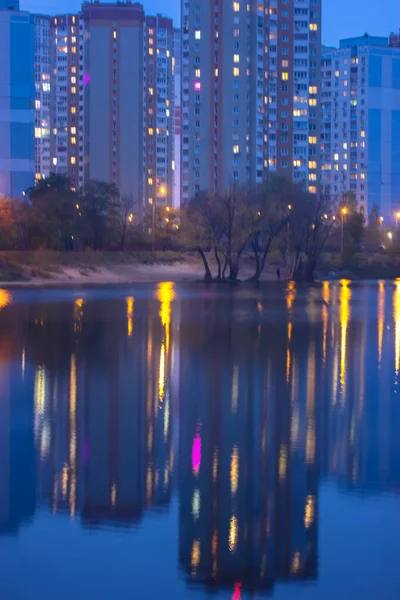 Des silhouettes de maisons à plusieurs étages avec une lumière vive dans les fenêtres se reflètent sur la surface de l'eau bleue dans le lac. Grande ville en détail — Photo
