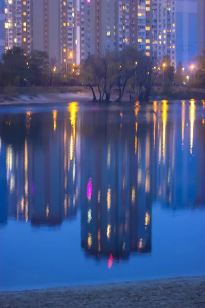 Silhuetas de casas de vários andares com luz brilhante nas janelas refletem sobre a superfície da água azul no lago. Cidade grande em detalhes — Fotografia de Stock