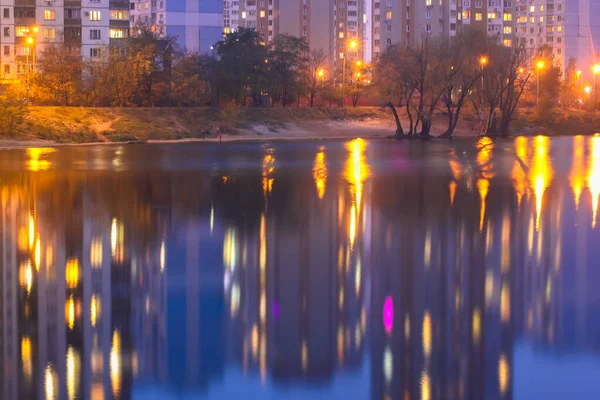 Silhuetas de casas de vários andares com luz brilhante nas janelas refletem sobre a superfície da água azul no lago. Cidade grande em detalhes — Fotografia de Stock