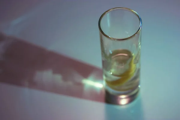 Vodka ou gim em um copo de tiro com uma fatia de limão dentro na superfície da mesa branca. Fundo azul e sombra de contraste — Fotografia de Stock