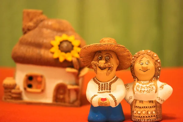 Husband and wife in national Ukrainian costumes on the background of a white clay house and blooming sunflower, miniature figures molded from clay