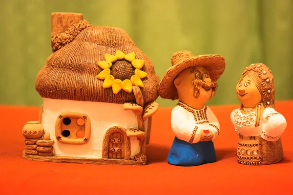 Toy husband and wife in national Ukrainian costumes on the background of a white clay house and blooming sunflower, miniature figures molded from clay