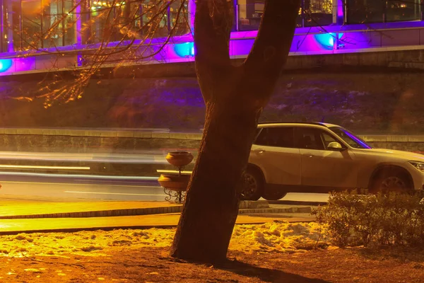 Carro branco SUV e tronco de árvore em primeiro plano. O movimento de carros na estrada no escuro, exposição longa — Fotografia de Stock