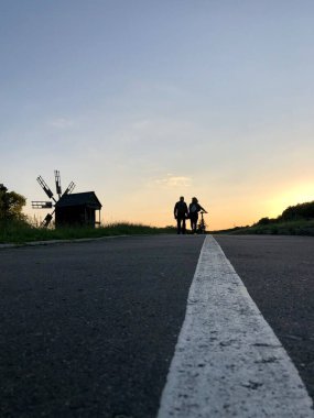 Bisiklet yolu ve iki bisikletçi gün batımında, ufuktaki rüzgarlı ahşap yel değirmeninin silüeti ile uzaklığa doğru yürür. Pirogovo, Kiev, Ukrayna