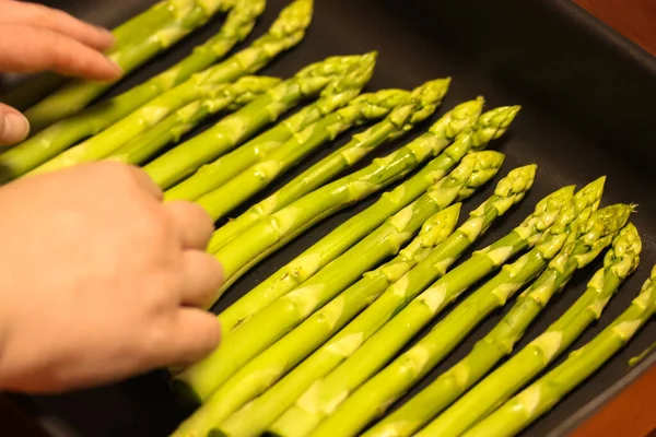 Womens hands put bundle of fresh organic asparagus on a black baking tray, cooking vegan food at home — Stock Photo, Image