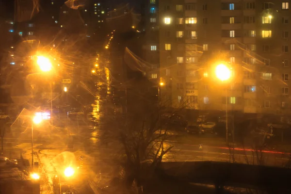 Rain outside the window in the city at night. Blurred lights of streetlights and fr cars driving down the road in bad weather. View from the window. High quality photo