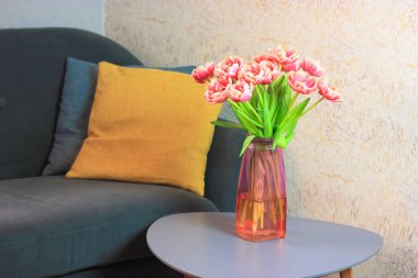 İçi rahat oturma odası, gri kanepe ve üzerinde sarı yastık. Küçük gri masadaki pembe cam vazoda pembe ve beyaz laleler..
