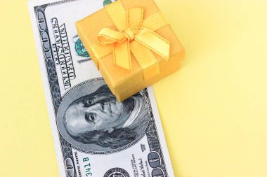 Soluk sarı arka planda 100 dolarlık banknot ve sarı mücevher hediye kutusu. Tatillerde pahalı hediyeler. Üst Manzara. Dümdüz. Metin için yer.