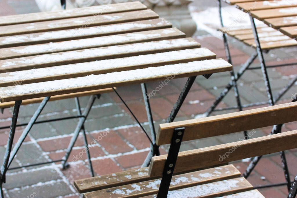 Wooden brown folding chairs and a table sprinkled with snow. Garden furniture on the terrace of the cafe in winter. Top view of empty chairs tables of street cafe in a big city. Selective focus.