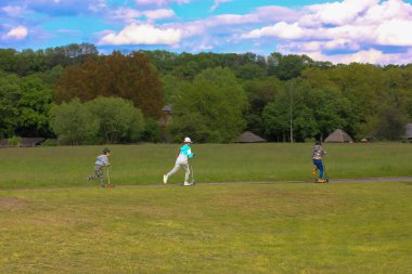 Tanımlanamayan çocuklar güneşli ve sıcak bir günde parktaki bir pistte scooterla geziyorlar. Okul çocukları için bahar tatili eğlencesi, aktif eğlence. Yeşil çayırlar ve yemyeşil ağaç taçları.