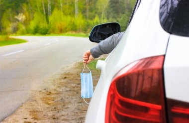 Beyaz bir SUV sürücüsü kullanılmış bir tıbbi maskeyi pencereden dışarı atıyor. Koronavirüs salgınının sonu. Arabayla seyahat etme kavramı. Normal hayata geri döndüm. Ormanda asfalt yol.