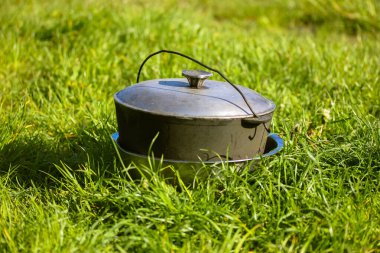 Güneşli bir yaz gününde yeşil çimlerin üzerinde gri alüminyum bir kazan duruyor. Kamp gezisi sırasında doğada yemek pişirmek. Seyahat, turizm, piknik pişirme, dökme demir kazanda kazıkta yemek pişirme..