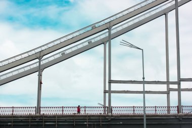 Gündüz vakti mavi bulutlu bir gökyüzüne karşı bir fener ve destekleyici yapısı olan modern bir yaya demirinden yapılmış köprü silueti. Kırmızı ceketli biri köprüde yürüyor. Şehir mimarisi.