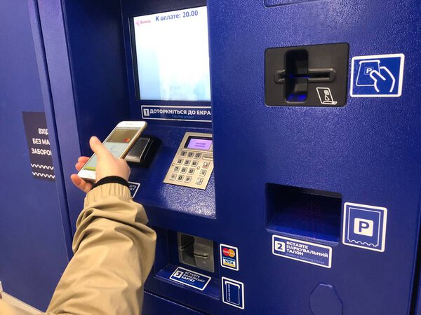 A driver paying parking fee in pay station, ATM paid parking. A person using terminal and smartphone with PayPass. A hand inserting ticket in the slot of a modern parking meter. Automatic parking time