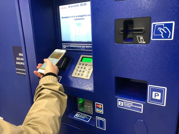 A driver paying parking fee in pay station, ATM paid parking. A person using terminal and smartphone with PayPass. A hand inserting ticket in the slot of a modern parking meter. Automatic parking time