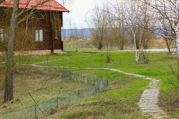 Route sinueuse pavée, sentier menant à la maison privée. Un chalet moderne en bois avec toit en tuiles rouges. Arbres nus et herbe verte sur une propriété au printemps, saison automnale. Espace privé. Immobilier. — Photo