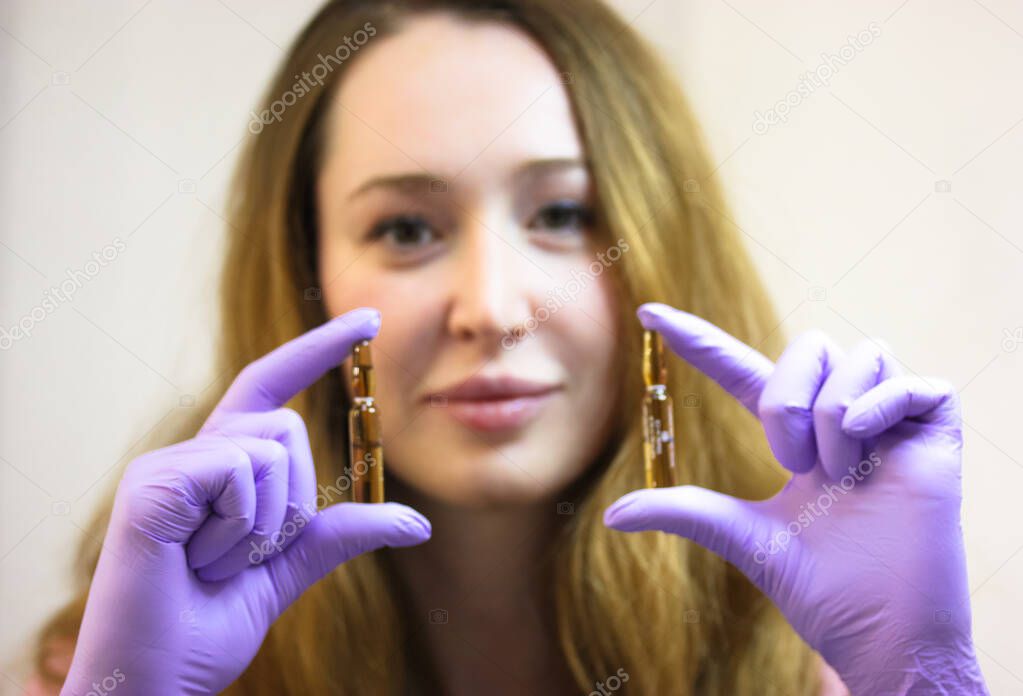 A young smiling cosmetologist in purple rubber gloves is holding ampoules of lifting serum. A concentrate in ampoules for correcting wrinkles, restoring facial skin elasticity. Skincare, cosmetology. 