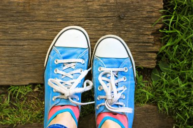 Mavi spor ayakkabılı bir kız tahta bir köprüde duruyor. Üst manzara, yeşil çimenlerin üzerinde kadın ayağı, güneşli yaz gününde ahşap zemin. Doğal bir manzara. Bir çift klasik klasik ayakkabı..