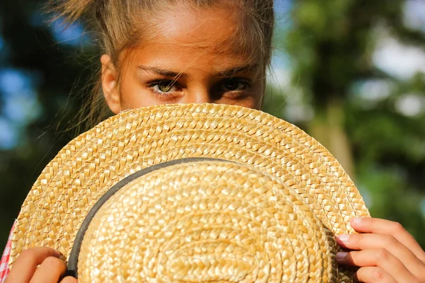 A beautiful little teenage green-eyed girl hides face behind a straw hat. A child hides half of her face behind a hat at sunny day. Summer vacation on nature, park, wood. Serious facial expression.