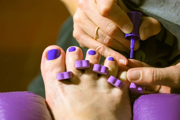 Pedicure master applies bright purple nail polish to client\'s nails in beauty salon. Women\'s fingers, toe divider up close. Pedicure Accessories. Girl applying nail varnish to toe nails at spa center.