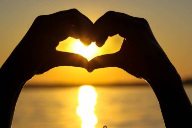 Gün batımında, sarı-turuncu bir gökyüzüne, güneş ışınlarına ve güneş ışığına karşı kalbinde bir kadın eli silueti. Aşkın sembolü. Deniz kenarında tatil, okyanus, göl, yazları nehir. Deniz kıyısı, sahil.