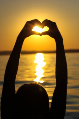 Gün batımında, sarı-turuncu bir gökyüzüne, güneş ışınlarına ve güneş ışığına karşı kalbinde bir kadın eli silueti. Aşkın sembolü. Deniz kenarında tatil, okyanus, göl, yazları nehir. Deniz kıyısı, sahil.