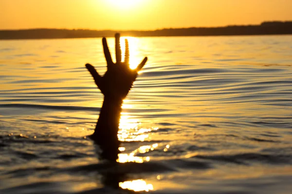 A man is drowning in water on a sunset. A hand peeks out from under a water. A drowning person needs help, rescue. A risk, danger for life in a sea, river, ocean. Death, SOS concept. Drowning victim.