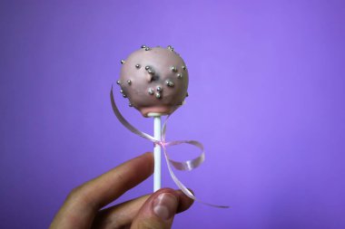 Pink chocolate cake pops with strawberry flavour on a purple background. A round cake on a stick decorated with ribbon and confectionery sprinkles of silver beads. A sweet treat. World Chocolate Day. clipart