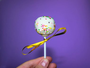 Mor bir arka planda beyaz çikolatalı kek beliriyor. Altın kurdeleyle süslenmiş yuvarlak bir çubuk üzerinde yuvarlak pasta, çok renkli boncuklar serpiştirilmiş şekerleme. Tatlı bir ikram. Dünyanın En Tatlı Günü.