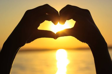 Gün batımında, sarı-turuncu bir gökyüzüne, güneş ışınlarına ve güneş ışığına karşı kalbinde bir kadın eli silueti. Aşkın sembolü. Deniz kenarında tatil, okyanus, göl, yazları nehir. Deniz kıyısı, sahil.