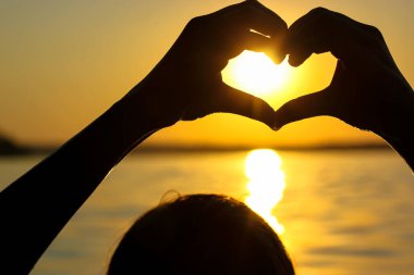 Gün batımında, sarı-turuncu bir gökyüzüne, güneş ışınlarına ve güneş ışığına karşı kalbinde bir kadın eli silueti. Aşkın sembolü. Deniz kenarında tatil, okyanus, göl, yazları nehir. Deniz kıyısı, sahil.