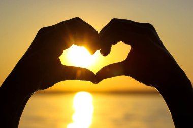 Gün batımında, sarı-turuncu bir gökyüzüne, güneş ışınlarına ve güneş ışığına karşı kalbinde bir kadın eli silueti. Aşkın sembolü. Deniz kenarında tatil, okyanus, göl, yazları nehir. Deniz kıyısı, sahil.