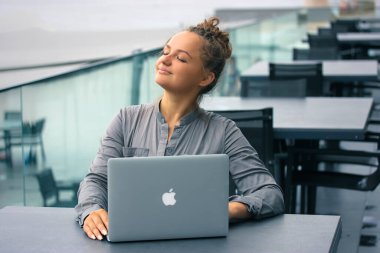 Kiev, Ukrayna, 17 Temmuz 2021. Gündüz vakti restoranın terasında Apple MacBook dizüstü bilgisayarı kullanan güzel bir iş kadını. Gri takımlı tatlı kız uzaktan çalışıyor, düşünüyor, hayal ediyor, gülümsüyor..