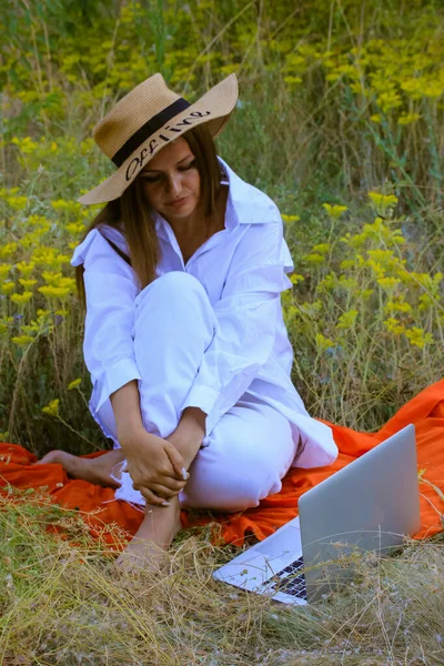 A young beautiful woman in the age 30 in a white suit, a straw hat with a text Offline sits in a summer blooming meadow and uses laptop. Concept of remote work, business online, vacation, recreation.