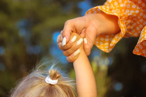 A young mother holds the infant girl\'s hand tightly, helping her take first step in life. A little girl with her hair gathered in a ponytail on a head at sunny summer day. Mother-daughter relationship