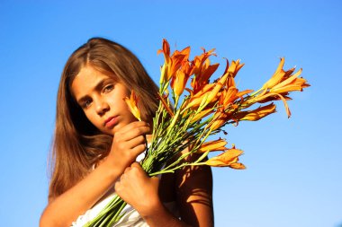Güneşli yaz gününde elinde turuncu zambaklarla mavi gökyüzüne bakan güzel bir genç kız. 10-12 yaşlarında bir kız çocuğu. Vahşi sarı çiçekler. 1 Eylül, okula dönüş, anneler günü konsepti..