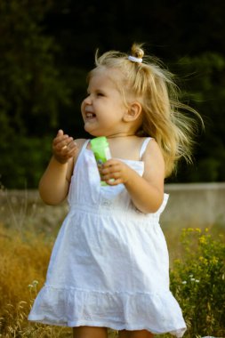 Dışarıda sabun köpüğü üfleyen mutlu, gülümseyen küçük bir bebek. 2-3 yaşlarında, beyaz elbiseli, yaz parkında yaşayan güzel sarışın bir çocuk. Çocuk eğleniyor, Joy. Pozitif duygular