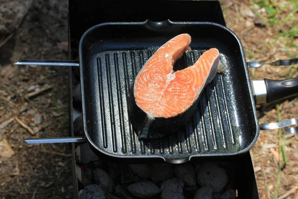 鮭のステーキを焼く 森の中のキャンプファイヤーの上の鉄板鍋で新鮮な口の中で生魚料理を散水赤 晴れた夏の日に田舎のピクニック 屋外ガスグリルで健康食品を準備します — ストック写真