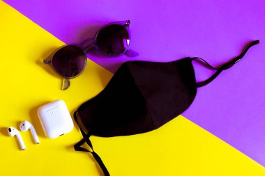 Siyah tıbbi yüz maskesi, mor güneş gözlükleri, sarı menekşe diyagonal arka planda kablosuz kulaklıklı kulaklıklar. Yeni normal. Salgın Covid 19. Şık aksesuarlar yaz tatili üst görünümü.