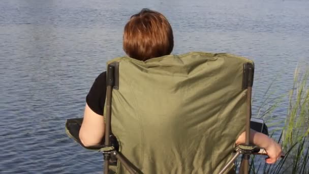 Uma mulher relaxa em uma cadeira de acampamento dobrável verde perto de um rio, observando um fluxo de água. Pesca no verão. A união dos humanos com a natureza. Um conceito de tranquilidade, harmonia, relaxamento, refrigeração ao ar livre. — Vídeo de Stock