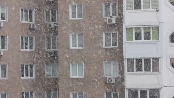 Vista da janela do apartamento na estação fria de inverno, vista dentro de casa. Grandes flocos de neve caindo lentamente ao ar livre durante o dia. Muitas janelas de uma casa de tijolo de edifício alto. Uma tempestade de neve na cidade. Vida urbana — Vídeo de Stock