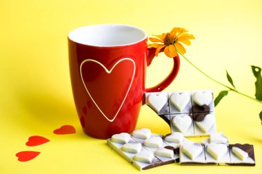 Kırmızı kupalı güzel bir kompozisyon, beyaz kalpli bir fincan, sarı çiçek, sarı arka planda kalpleri olan sütlü siyah çikolata. Sevgililer Günü tebrik kartı için harika bir şablon..
