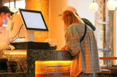 A cashier wearing a medical mask checkout, serves customers at fast-food during a pandemic. People in bistros, coffee shops study menus, order at a bar and pay for the food. POS monitor in cafeteria clipart