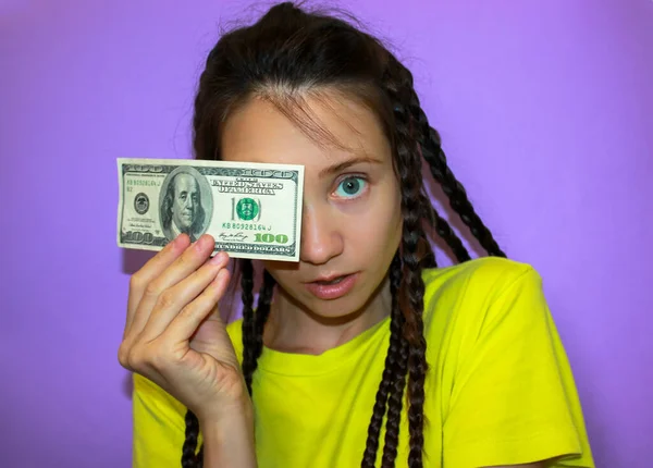 Surprised brunette blue-eyed girl in a yellow T-shirt on a purple background holding a hundred dollar note in hand, covering face. Success, business investment, finance, earning money, cash concept.