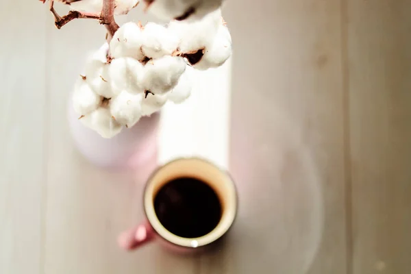 A cup of black coffee in a ceramic cup, mug in defocus, a branch of white cotton flower in a vase in focus on a white parquet floor background. Cozy home at autumn season. Sunlight. Place for text.